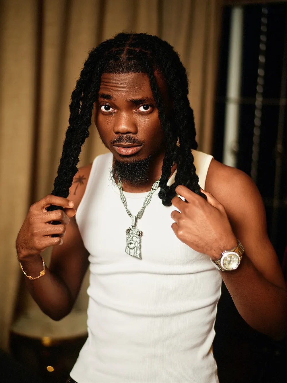 Emmy Blaq in a white vest and chains, holding his locs