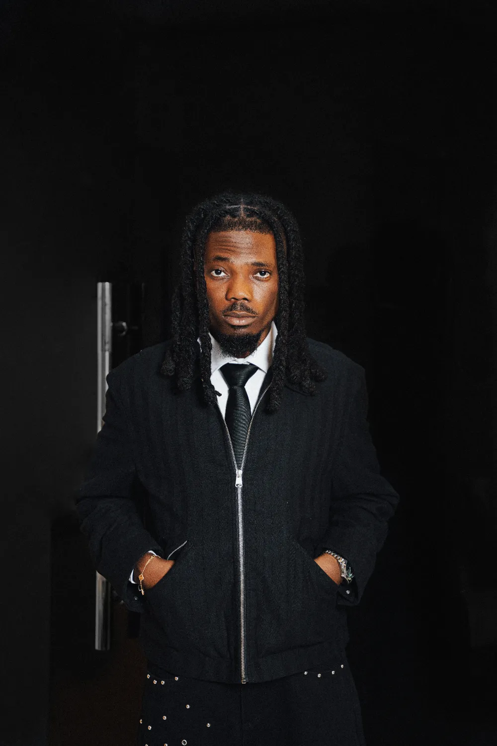 Emmy Blaq in a black pinstripe jacket and tie, standing in the dark