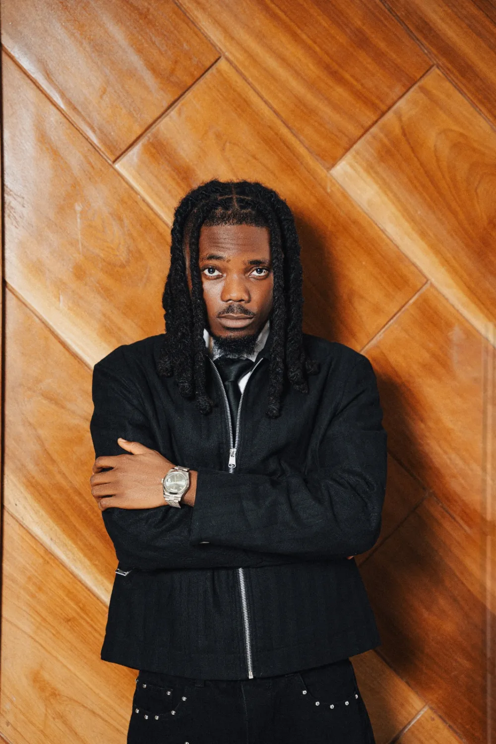 Emmy Blaq in a black zip jacket, arms folded, against a wooden wall