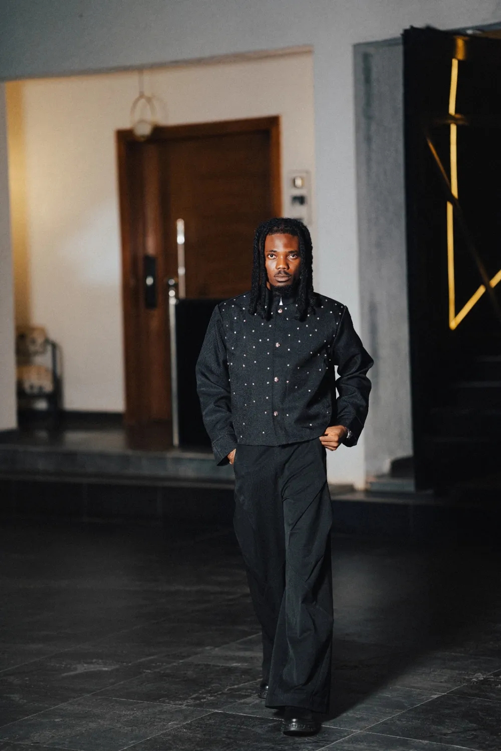 Emmy Blaq walking through a doorway in a studded black jacket and wide trousers